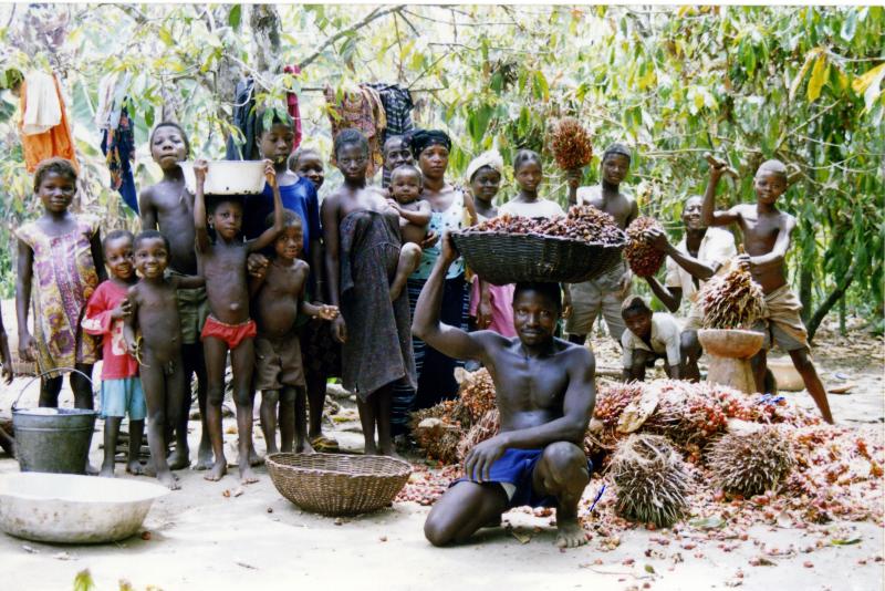 A group of children and adults gather in a lush outdoor setting, some carrying baskets of reddish fruits, with clothes hung on trees